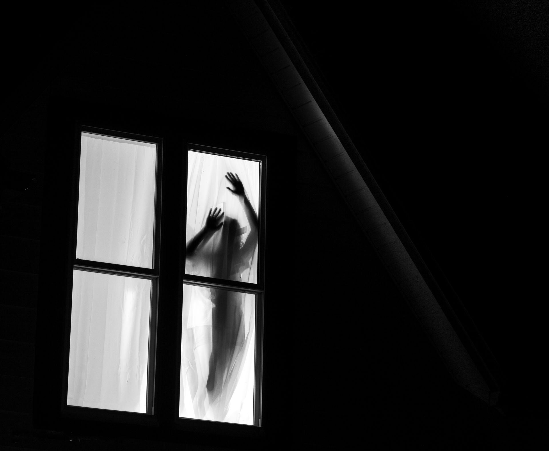Silhouette of a person with raised arms pressed against a window. Dark room, black and white.