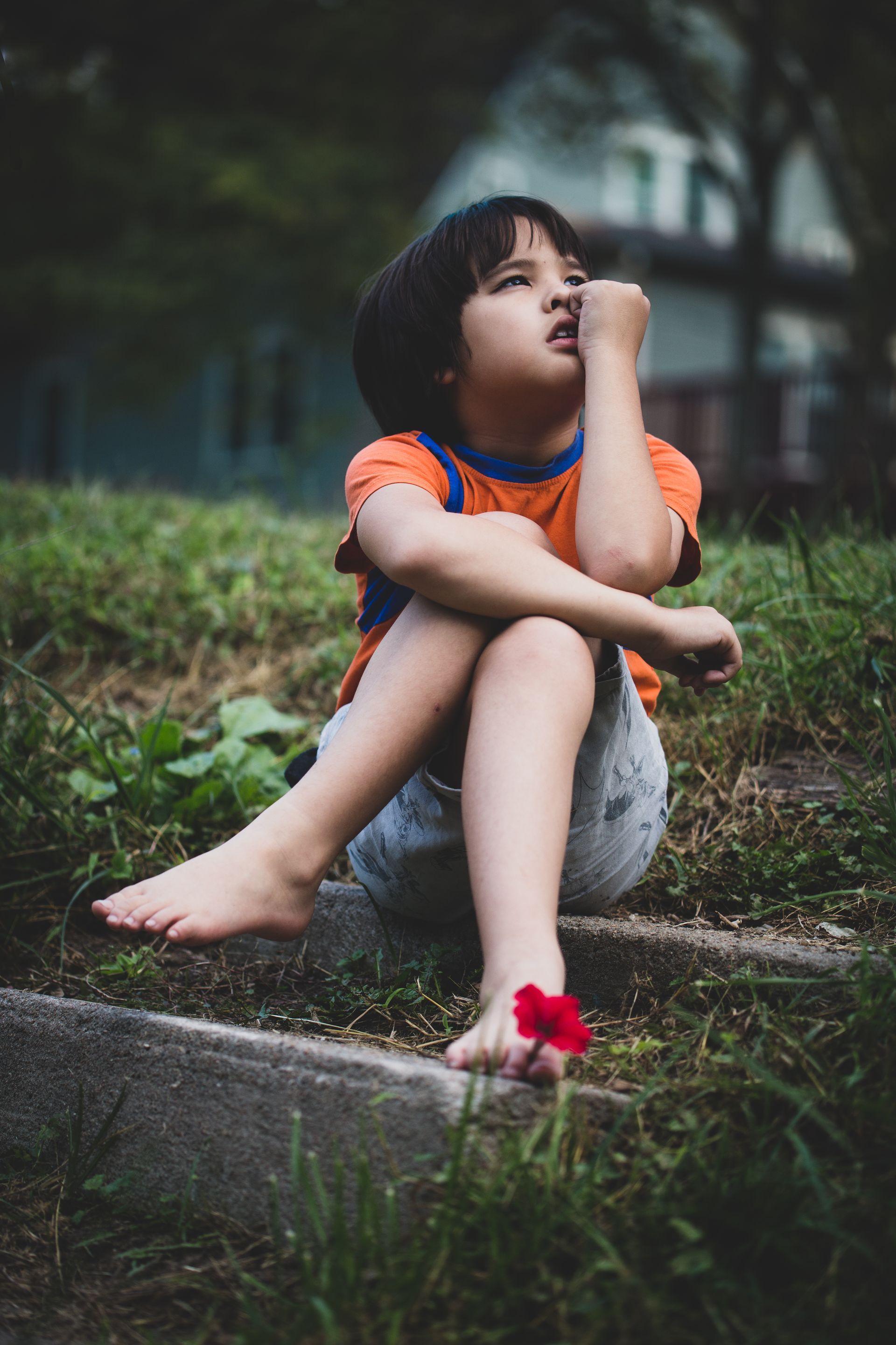 Boy sits on steps, looking up, bare feet with a red flower, grass, and a building in the background.