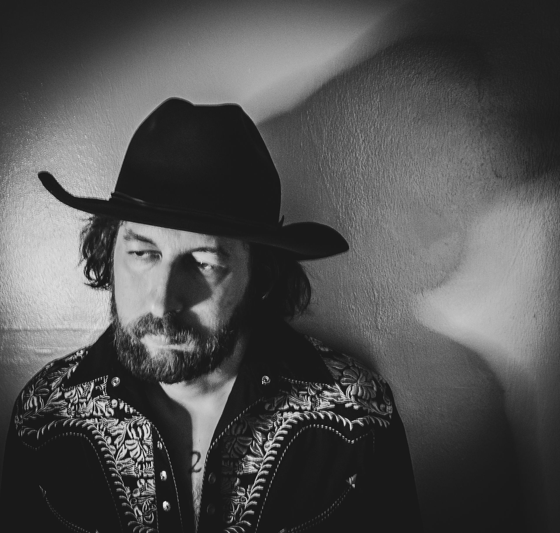 Man in cowboy hat, embroidered shirt, leans against a wall, black and white.