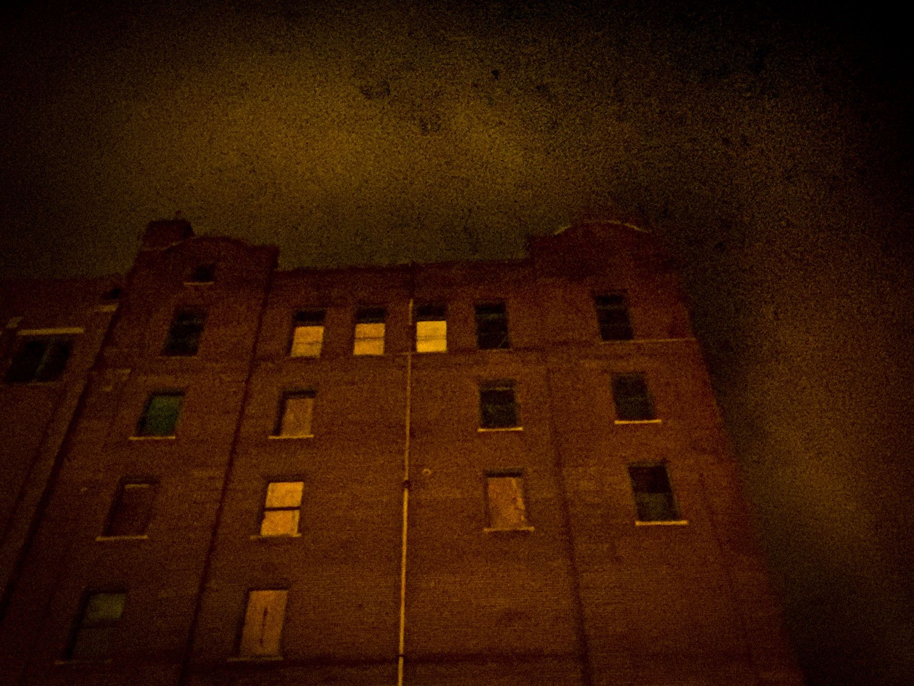 Dark, gritty photo of a brick building at night. Some windows lit from within, against a dark, textured sky.