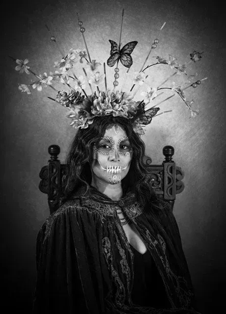 Woman in Day of the Dead makeup and floral crown, seated in antique chair, black and white.