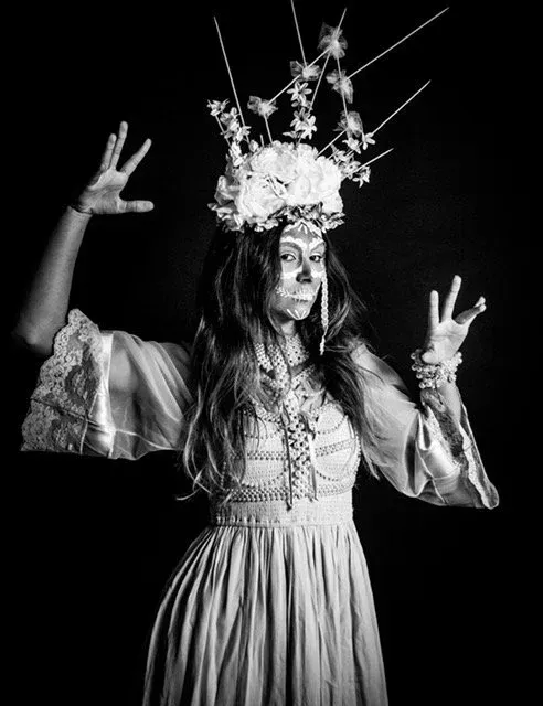 Woman in white dress and floral crown with arms raised, face painted, black background.