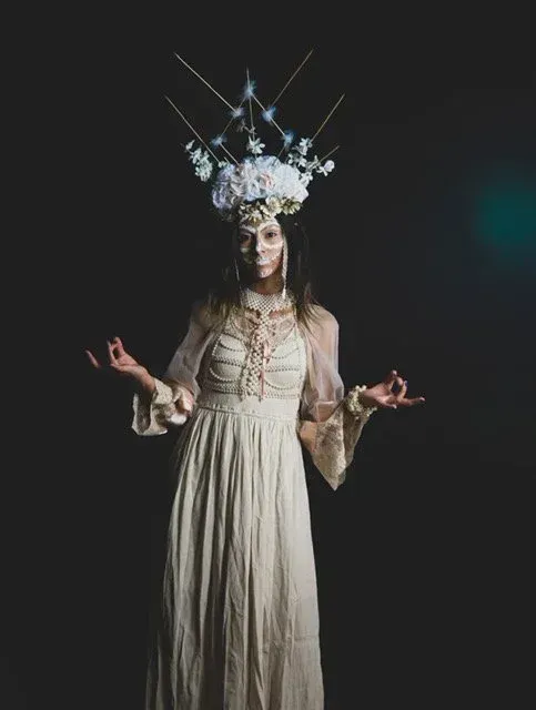 Woman in cream dress and floral crown, arms outstretched, face painted, against a dark backdrop.