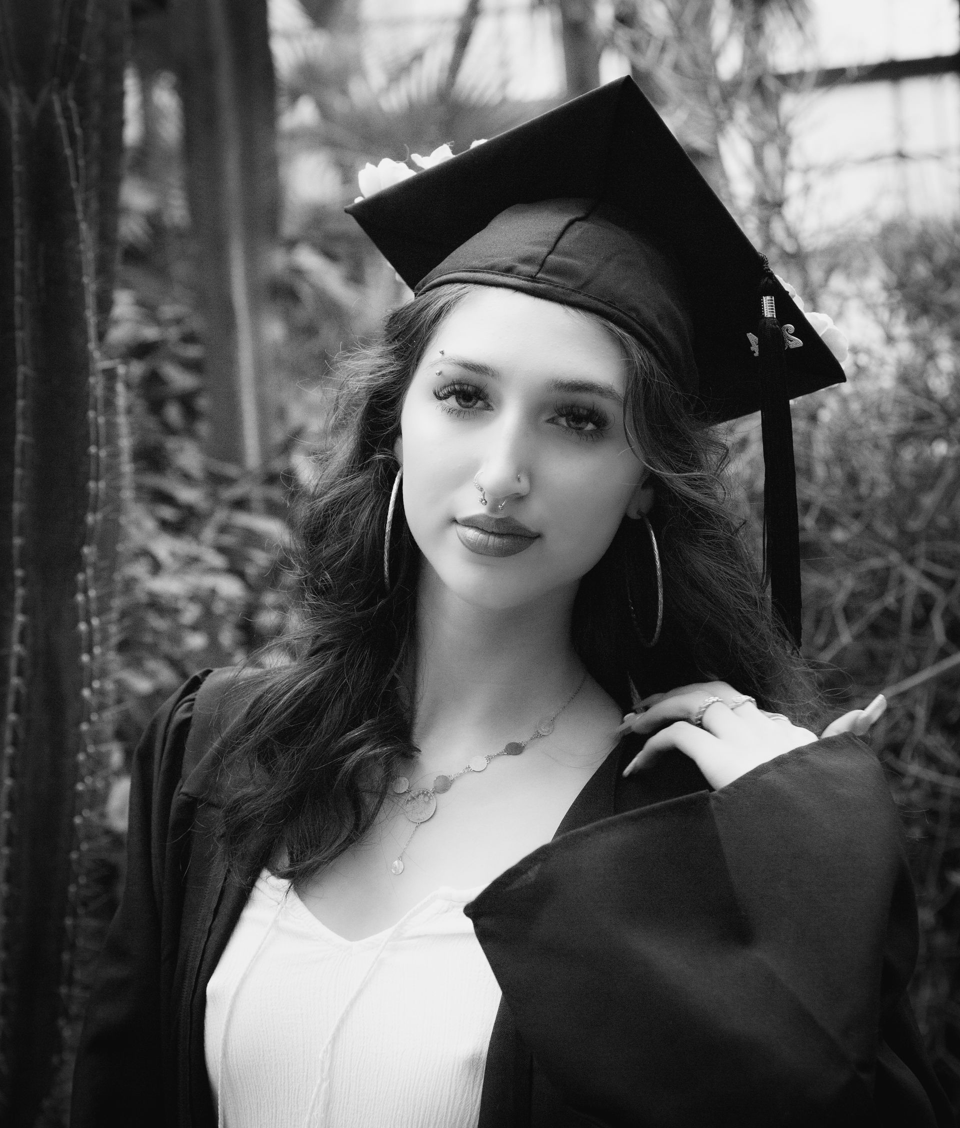 Young woman in graduation cap and gown smiles, in front of foliage.