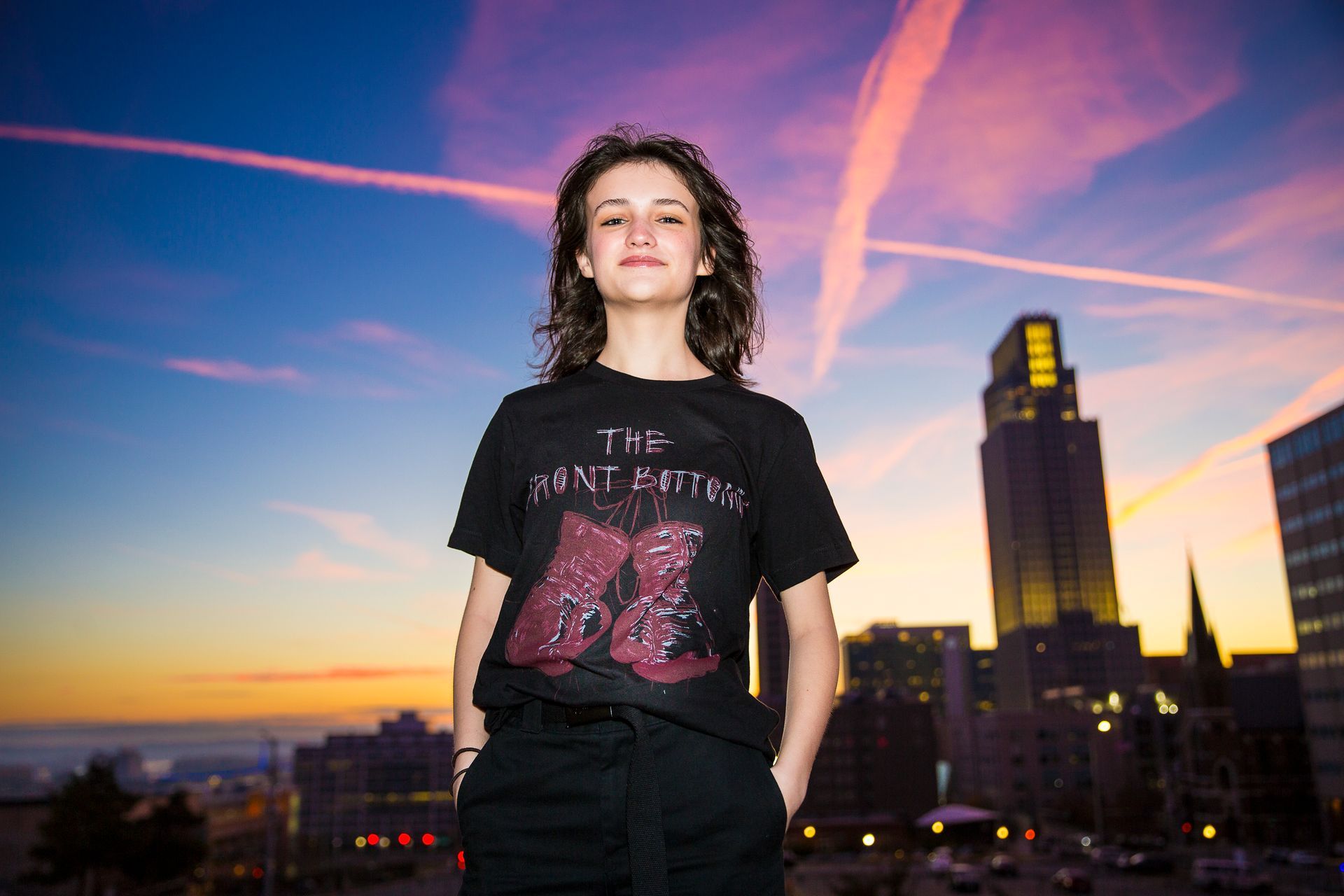 Woman in black shirt and pants with hands in pockets, posing in front of a sunset skyline.