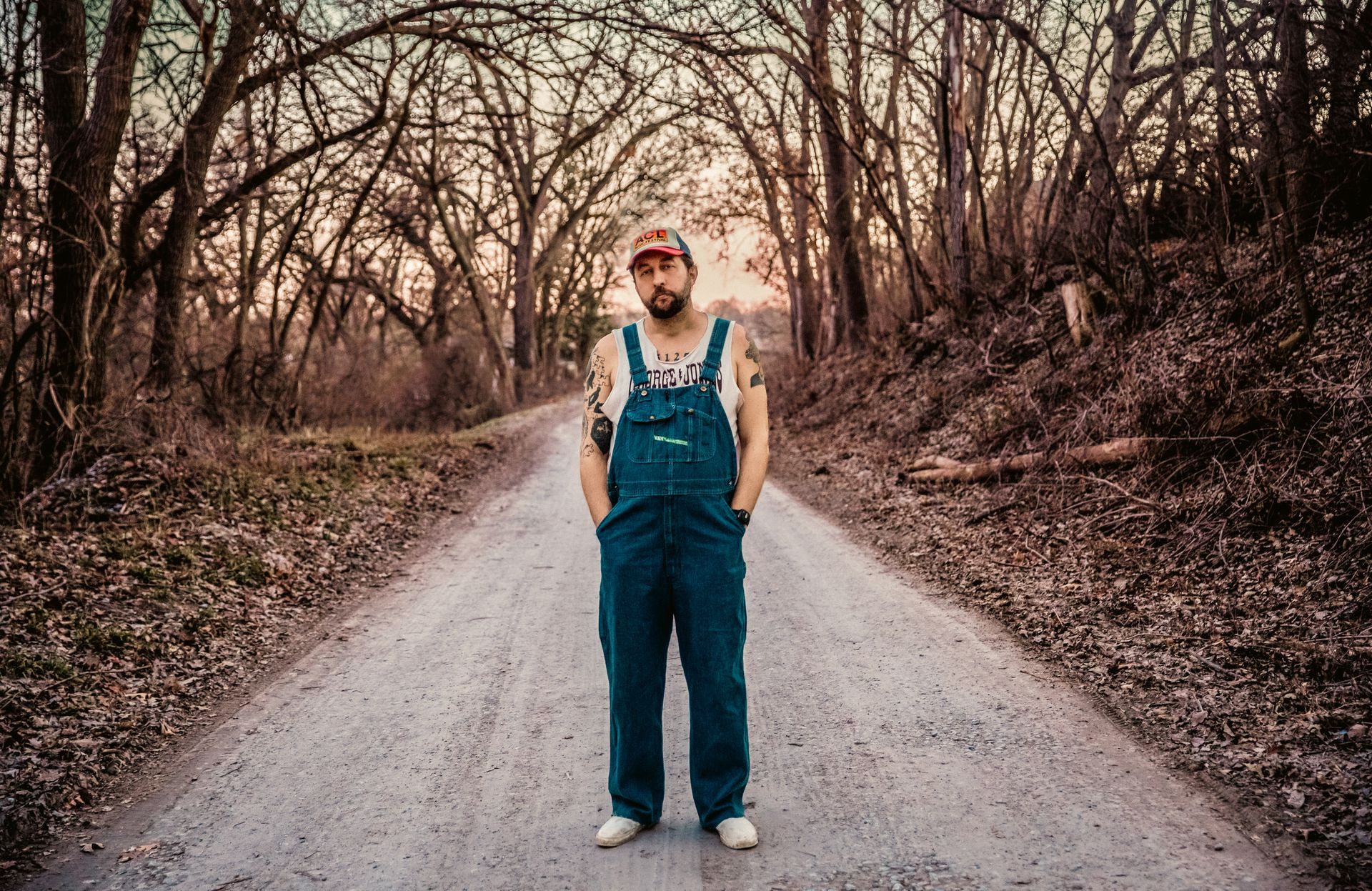 Man in overalls stands on a dirt road in a wooded area, hands in pockets.
