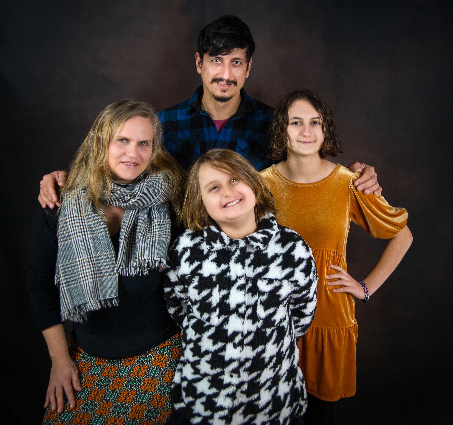 Family portrait: Two parents and two daughters pose against a brown backdrop. Smiling.