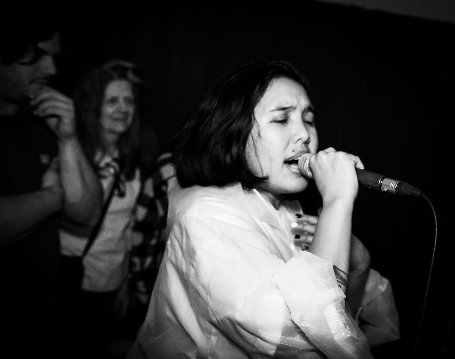 A woman sings into a microphone on stage, eyes closed. Two people stand in the background. Black and white.