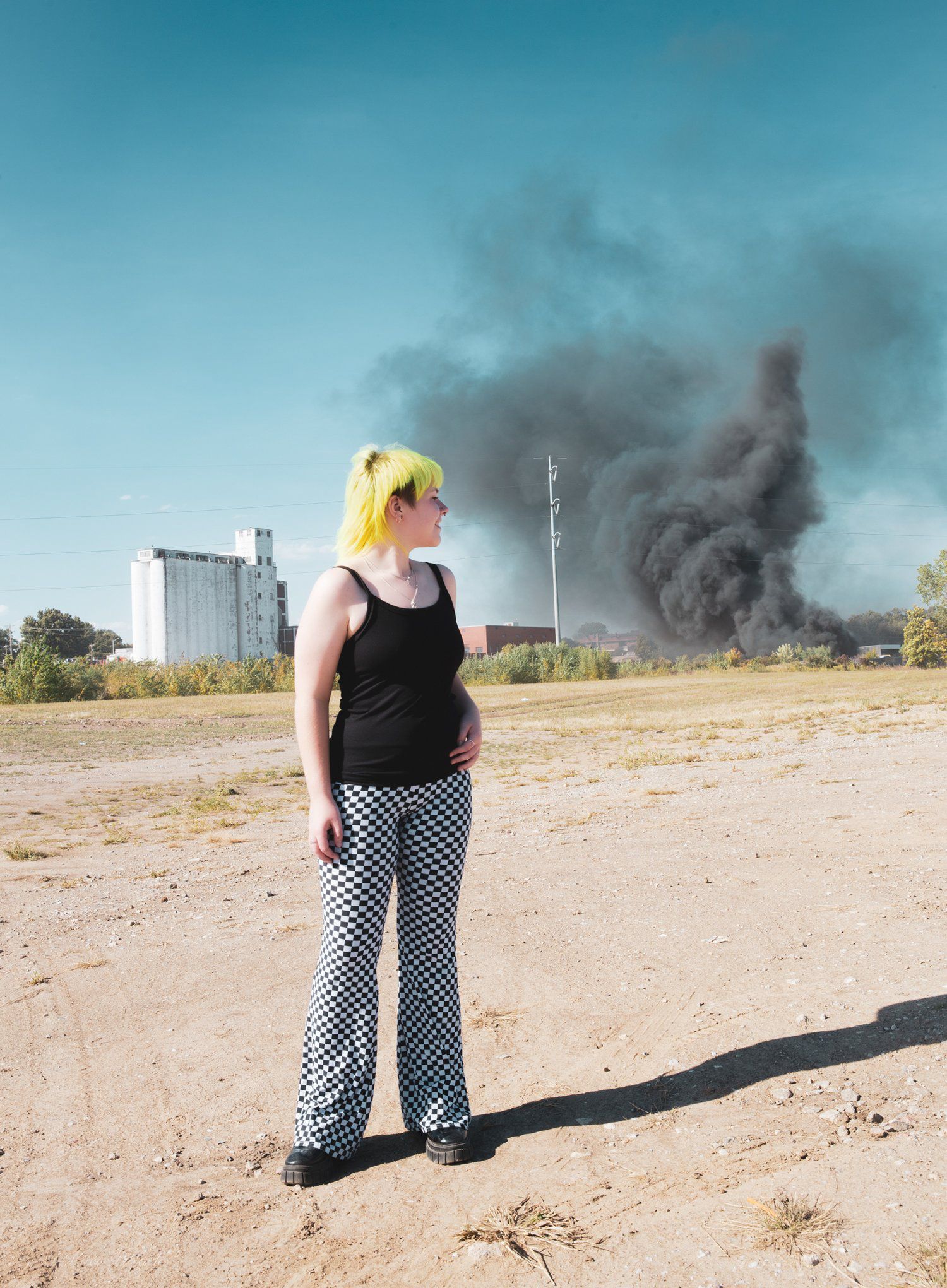 Woman with yellow hair looks at a large plume of black smoke. She stands on dirt, near a building.