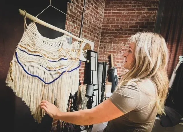 Woman examining a macrame wall hanging, cream and blue, hanging on a bamboo rod, brick wall backdrop.