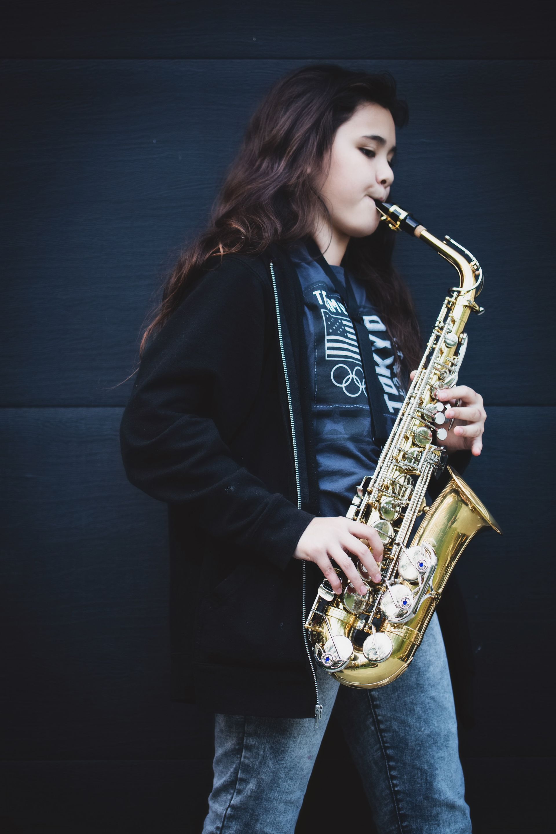 Young person with long hair playing a golden saxophone against a dark background.