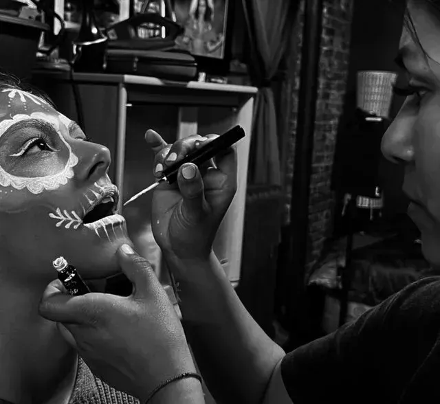 Person having Day of the Dead face makeup applied by another person, indoors. Black and white.