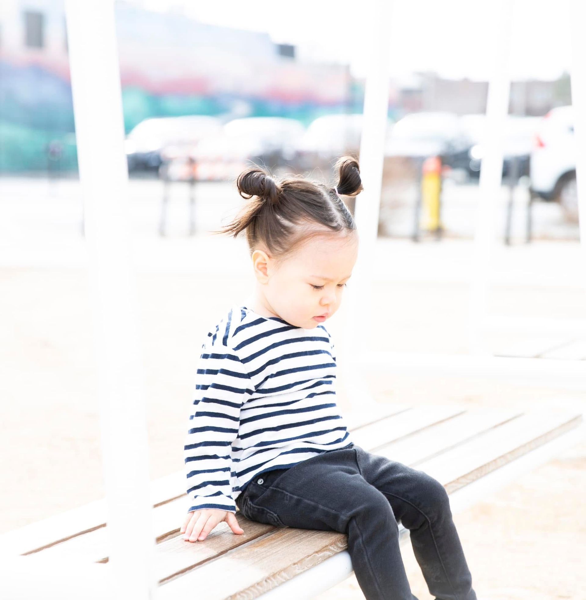 Young child with space buns, in striped shirt and black pants, sitting on a bench, looking down.