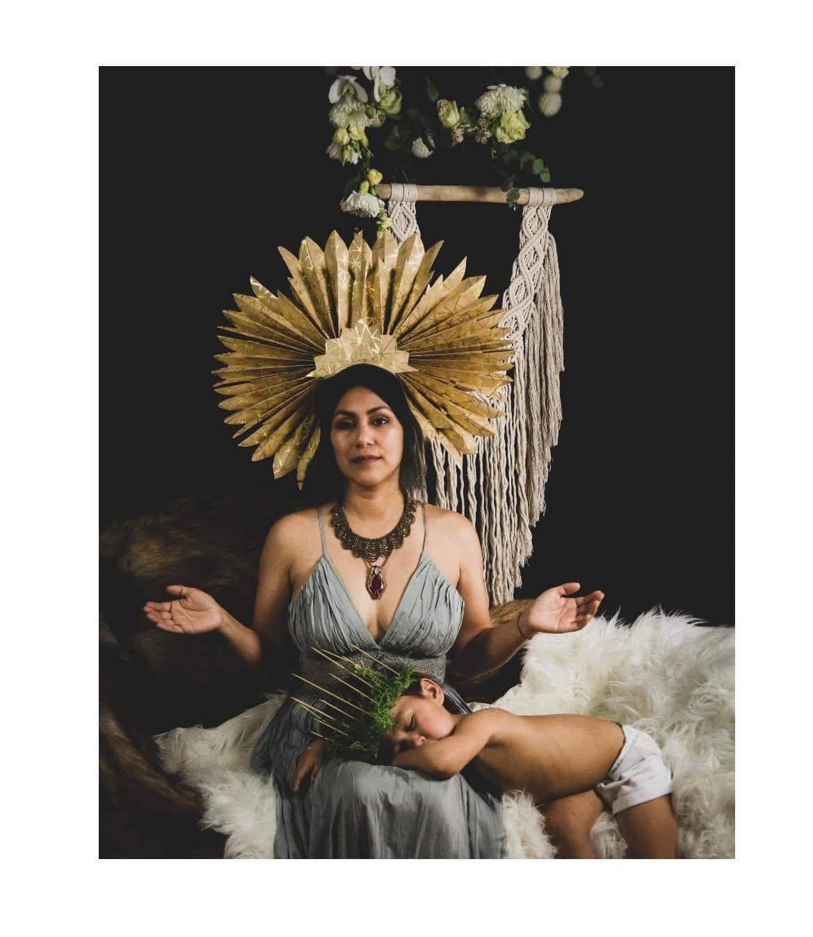 Woman with golden headpiece and child resting on her, both on a fur-covered surface, set against a black backdrop.