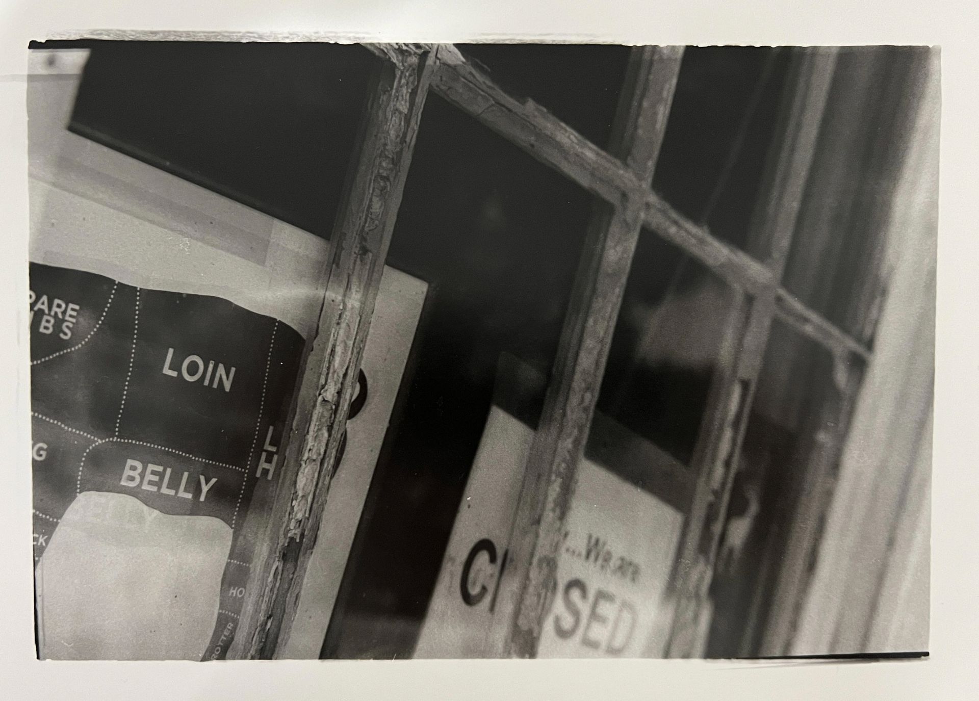 Black and white photo of a weathered shop window, meat cuts chart visible, 