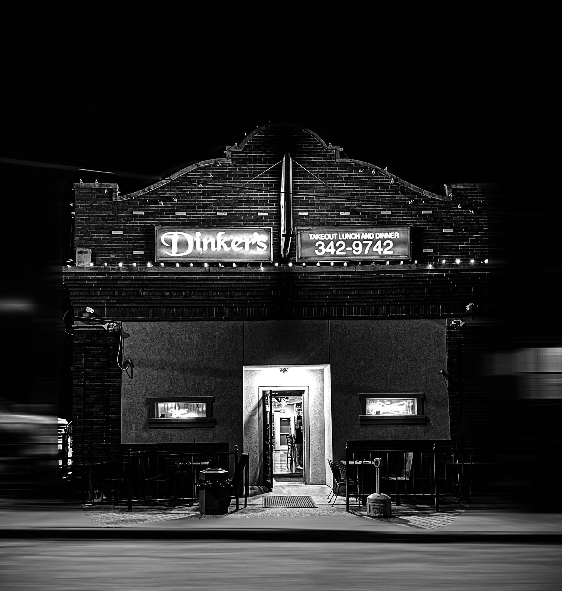 Black and white photo of Dinker's bar at night. Neon sign, brick building, open door, side windows.