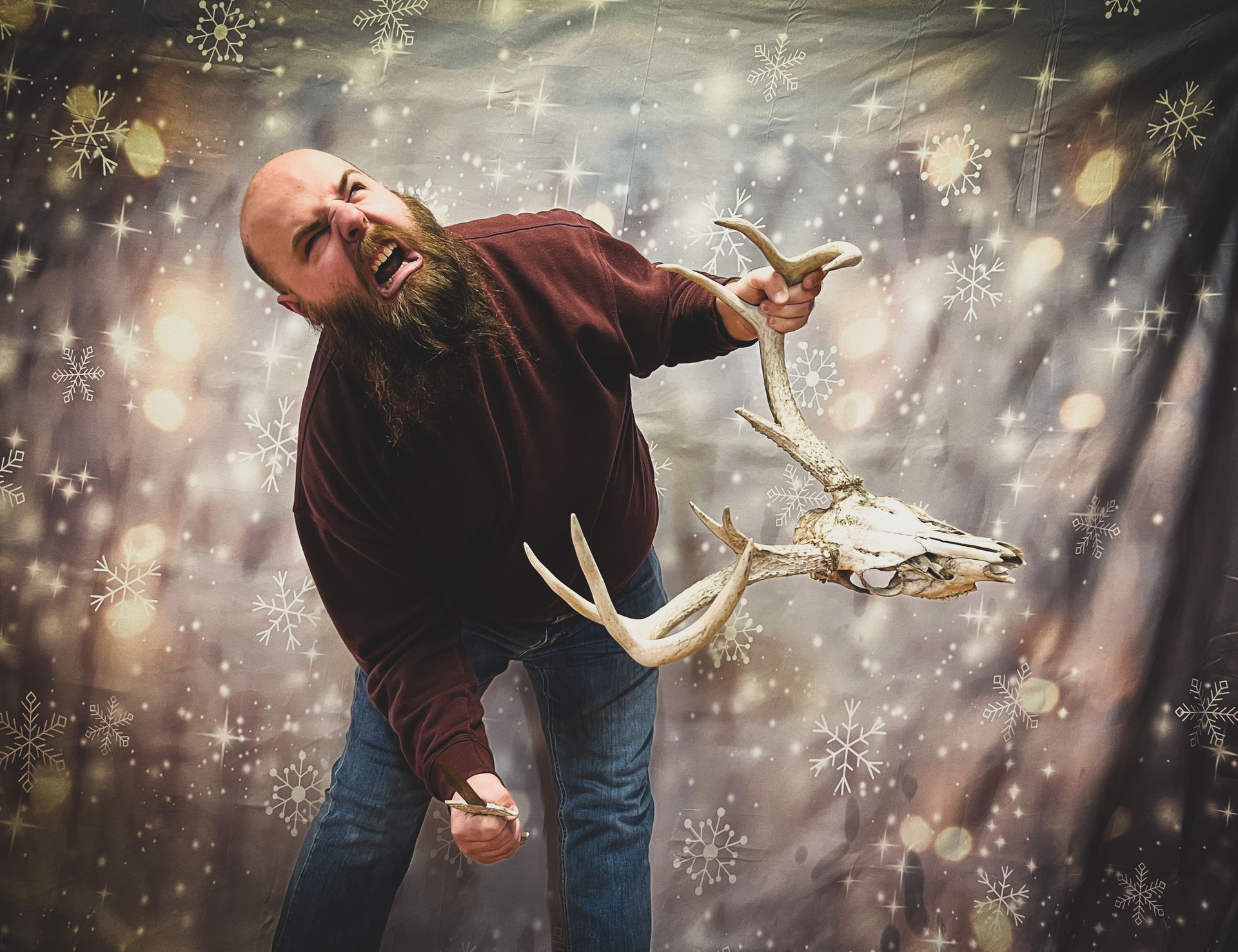 Bald man with a long beard, in a maroon shirt and jeans, struggles with a deer skull in front of a snowy backdrop.