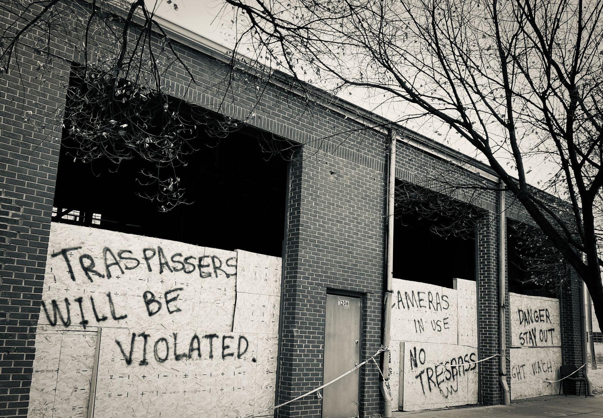 Black and white view of a boarded-up brick building with warning graffiti: 