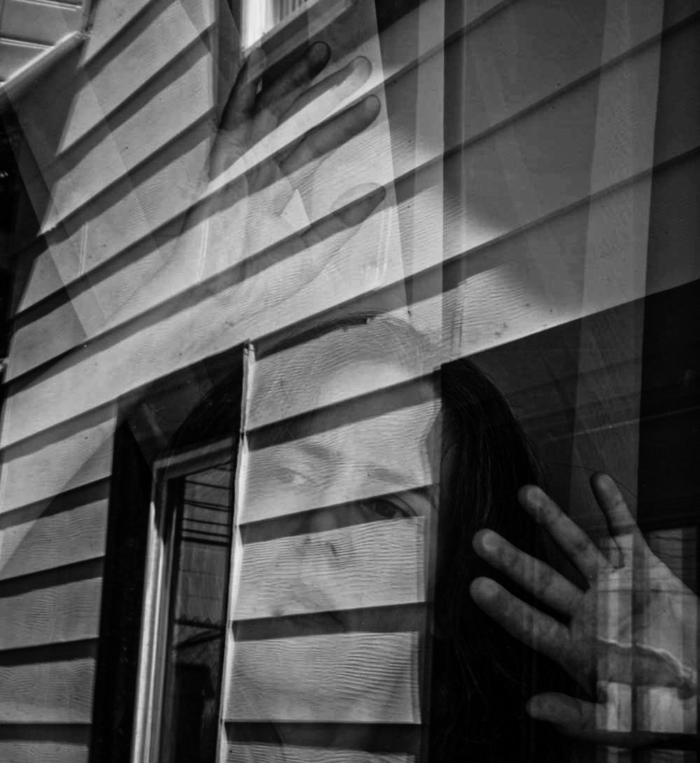 Black and white photo: hand presses against glass window reflecting a woman's face; horizontal siding on a building is visible.