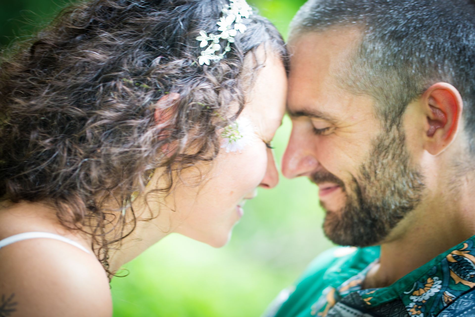 Couple touching foreheads, smiling, outdoors; woman with flowers in hair, man with beard, green background.