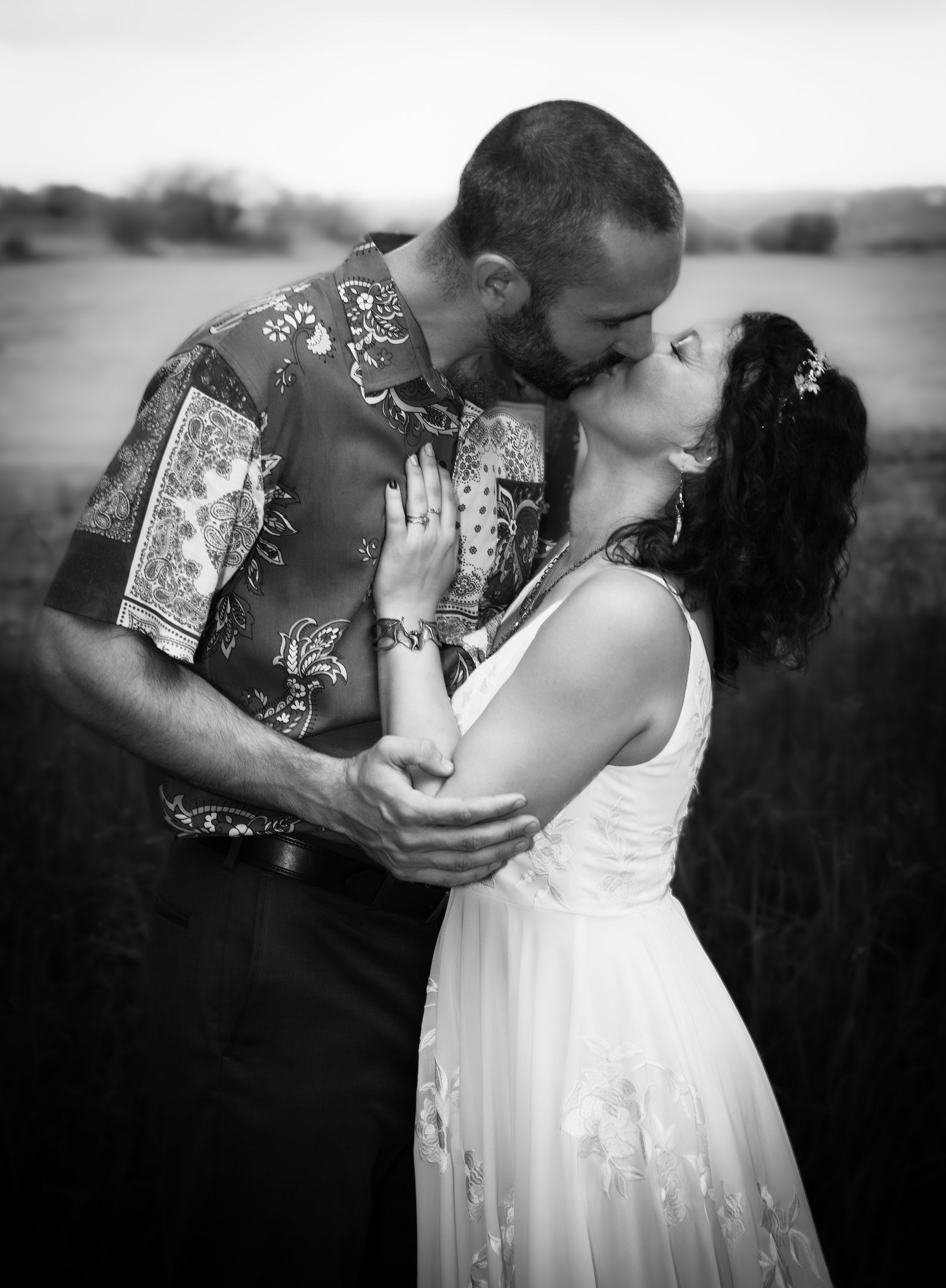 Couple kissing, the bride in a white dress and groom in a patterned shirt, in a field.