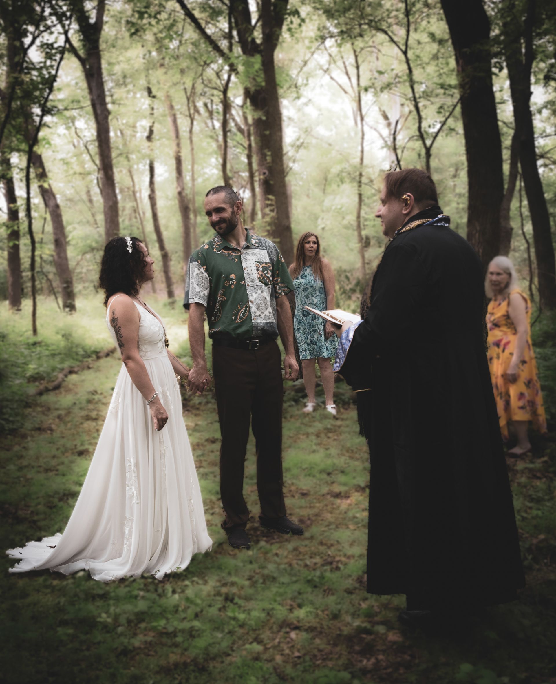 Couple exchanging vows in a forest, officiant in black robe. Guests in the background, trees surrounding.