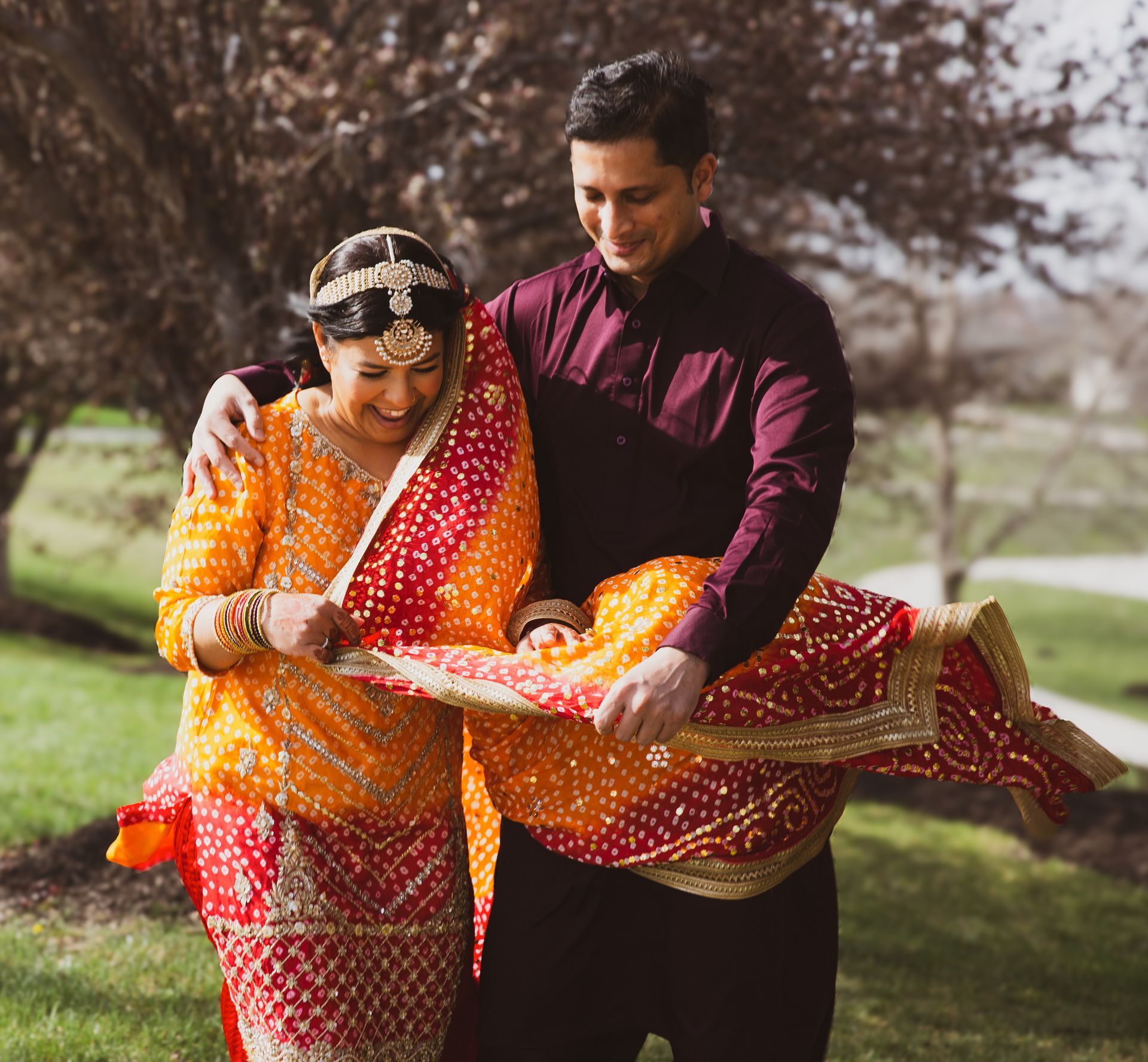 Man in burgundy shirt helps woman in yellow and red sari with dupatta, outdoors.