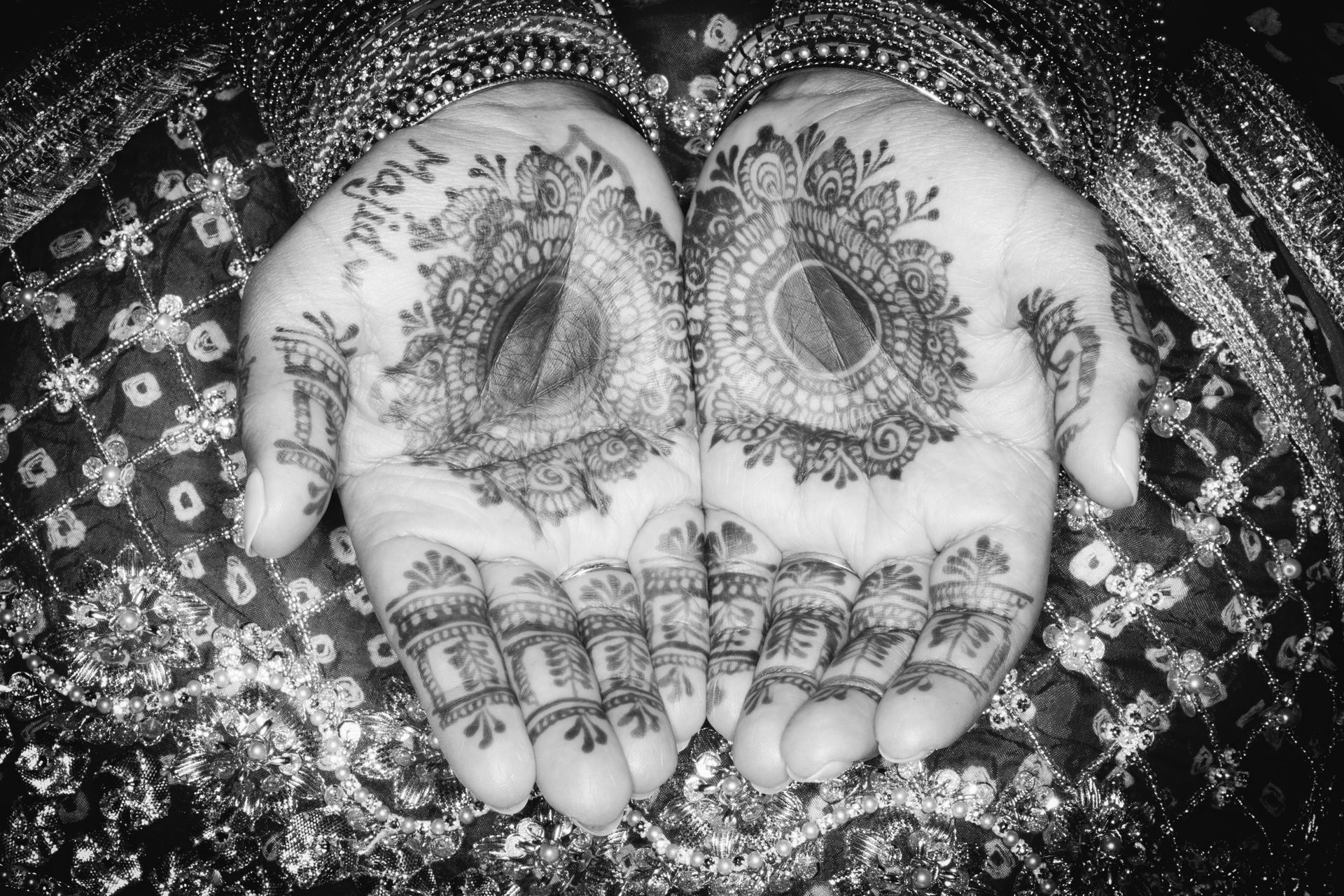 Black and white image of hands with intricate henna designs, likely for a wedding.