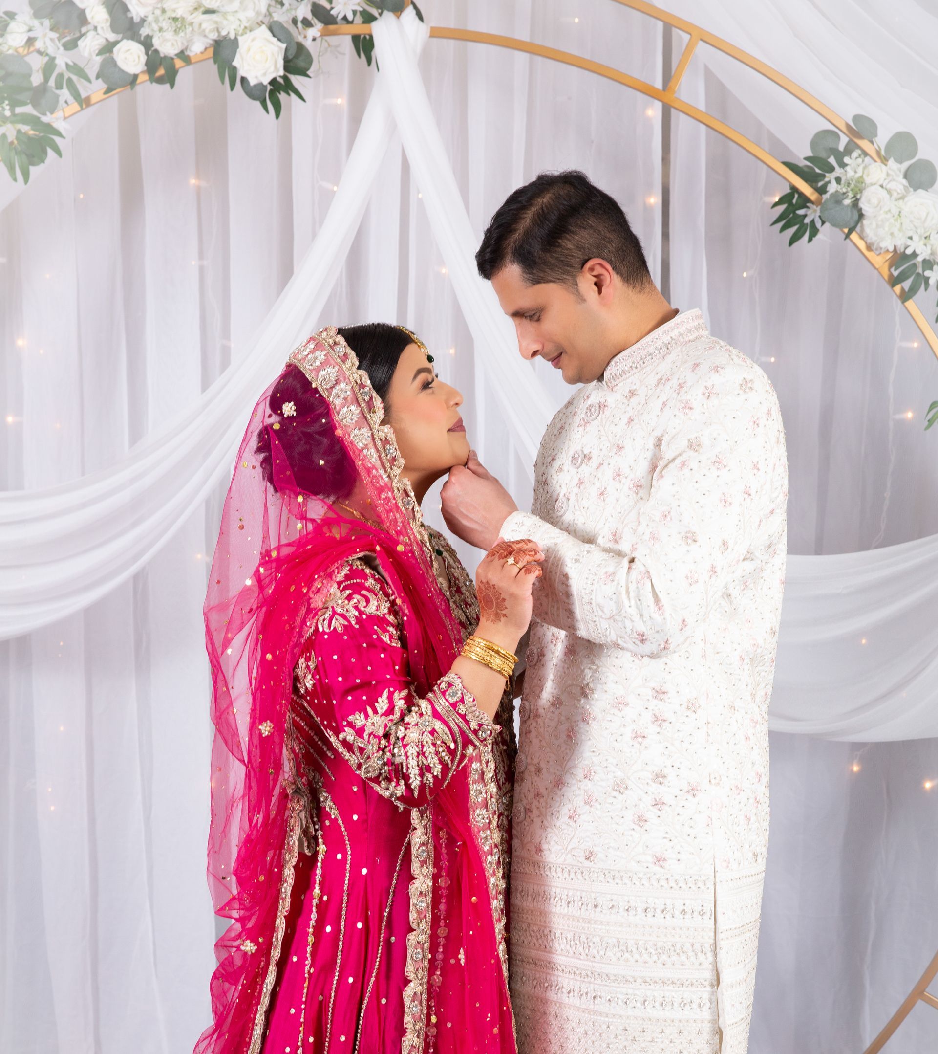 Couple in wedding attire, gazing at each other. Bride in pink, groom in white, under floral arch.