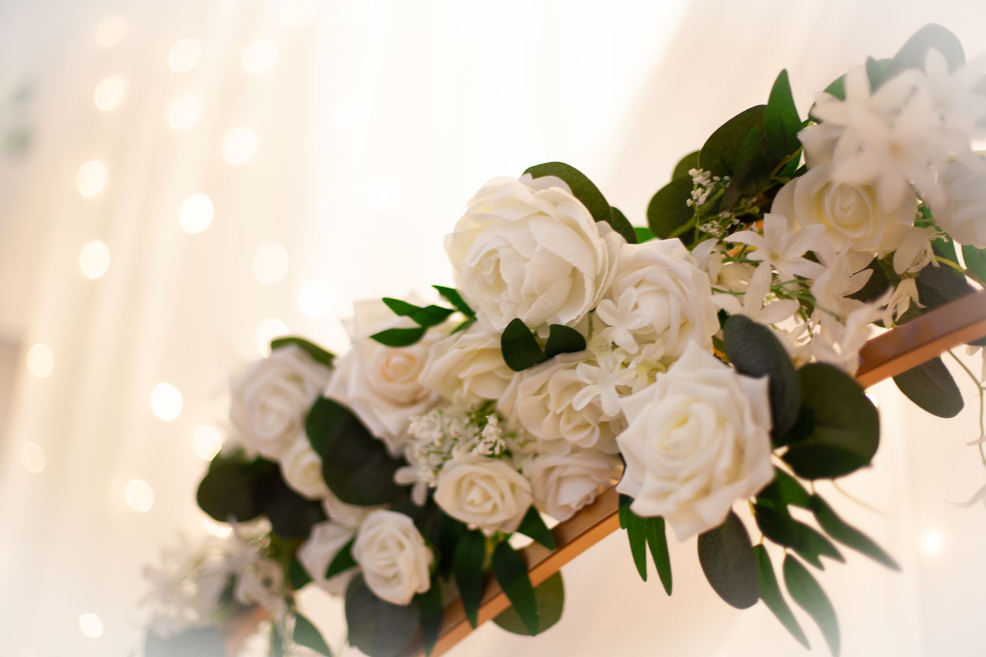 White roses and greenery decorate a wooden arch against a soft, blurred background of fairy lights.