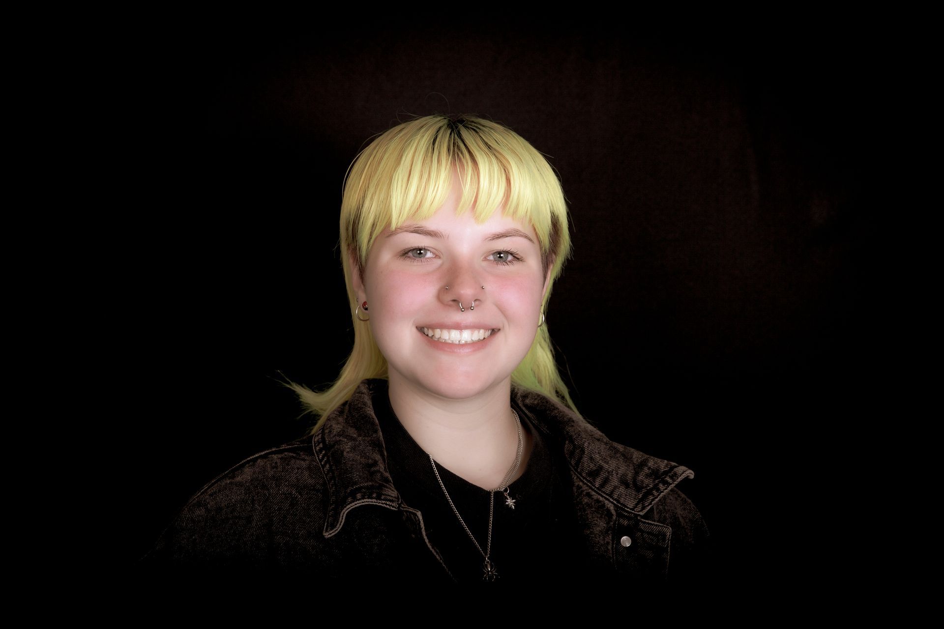 Person with light yellow hair, smiling, wearing a black jacket and a nose ring. Black background.