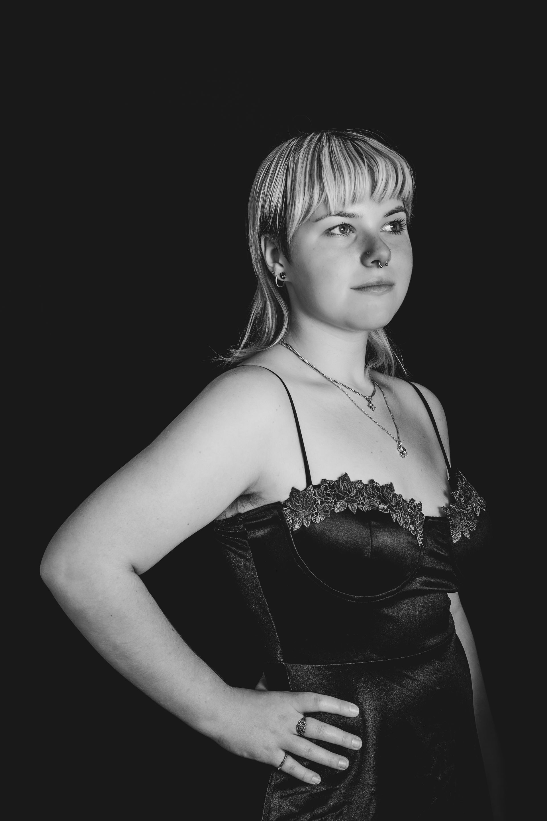 Woman in black dress, hand on hip, looking off to the side, black background.