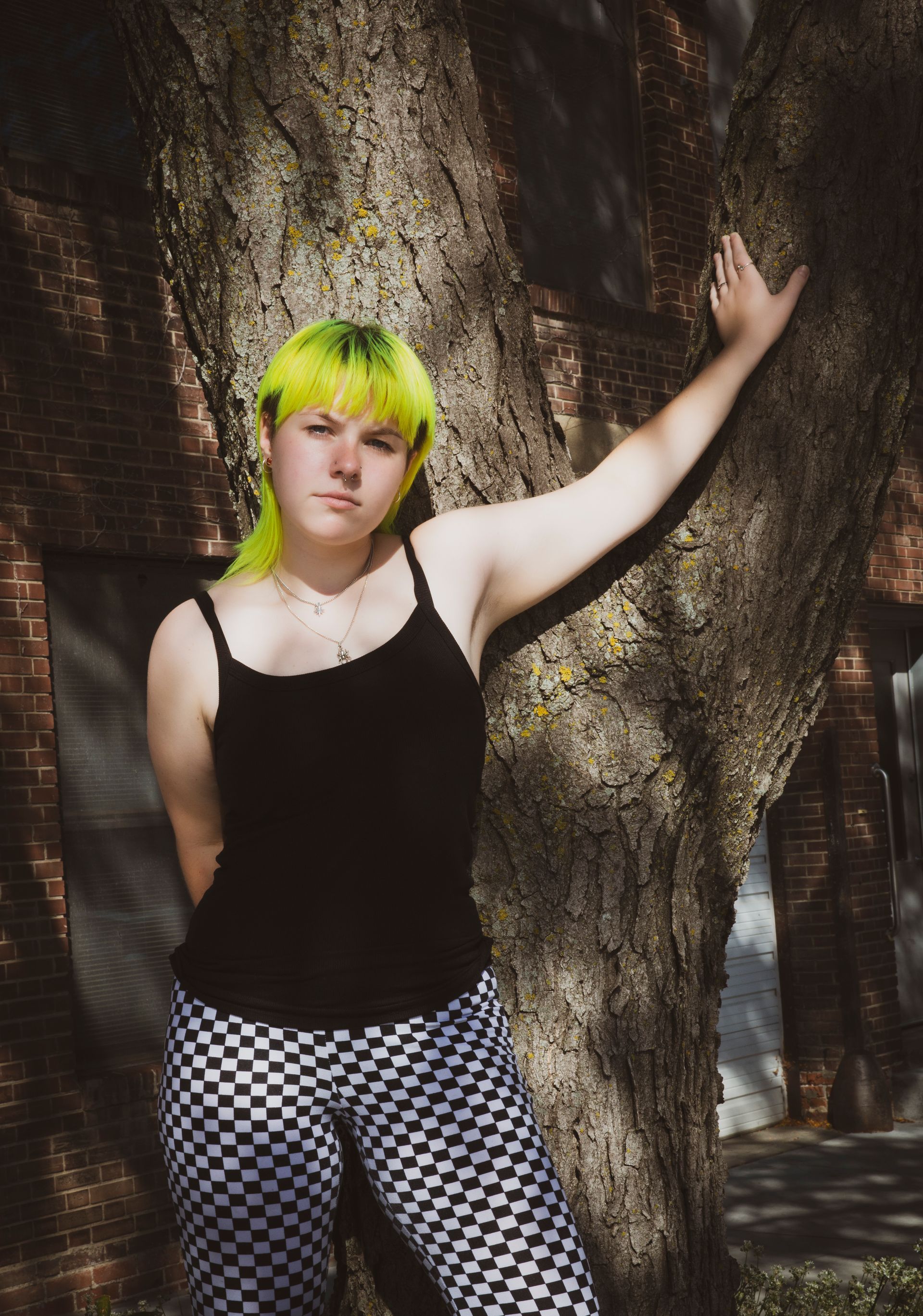 Person with bright green hair, black tank top, checkered pants, leaning against a tree, with an indifferent expression.