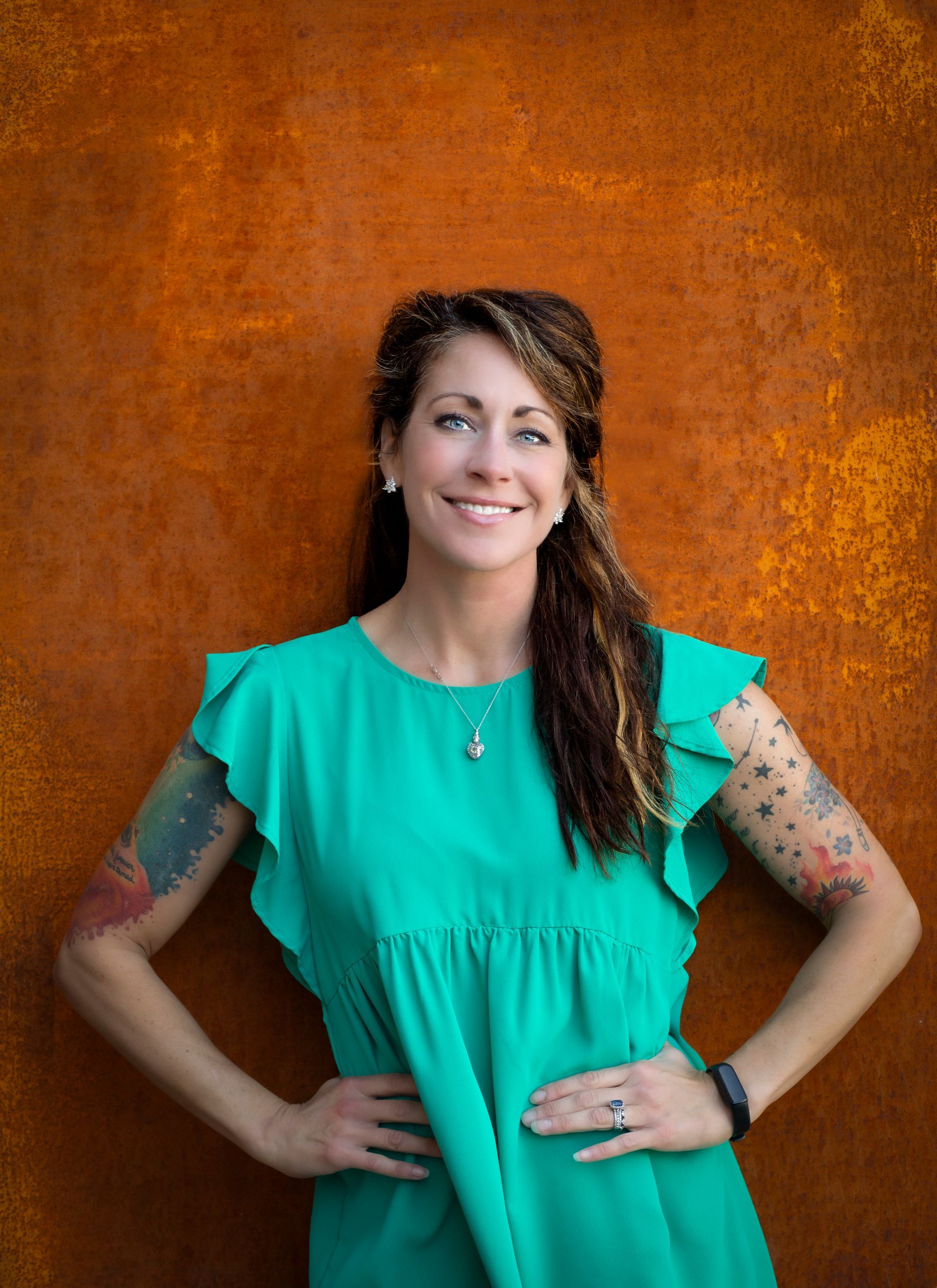 Woman with arm tattoos smiles, wearing teal dress, hands on hips, against rust-colored wall.