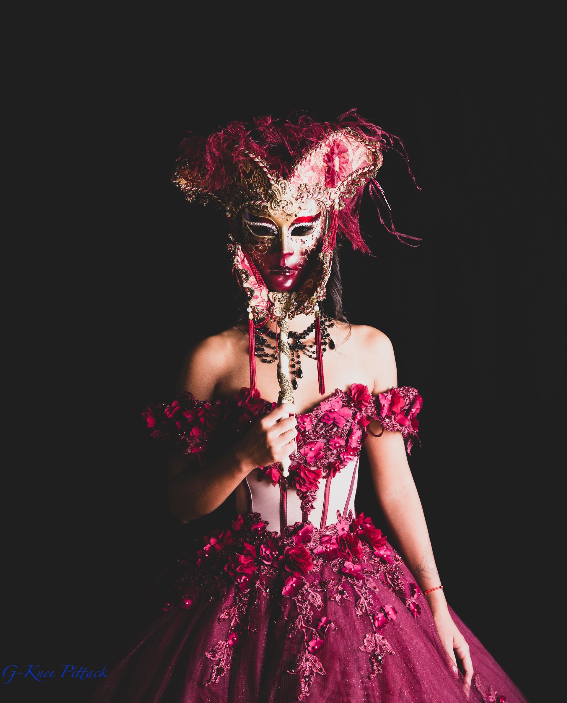 Woman in a burgundy ball gown and elaborate mask against a black background.