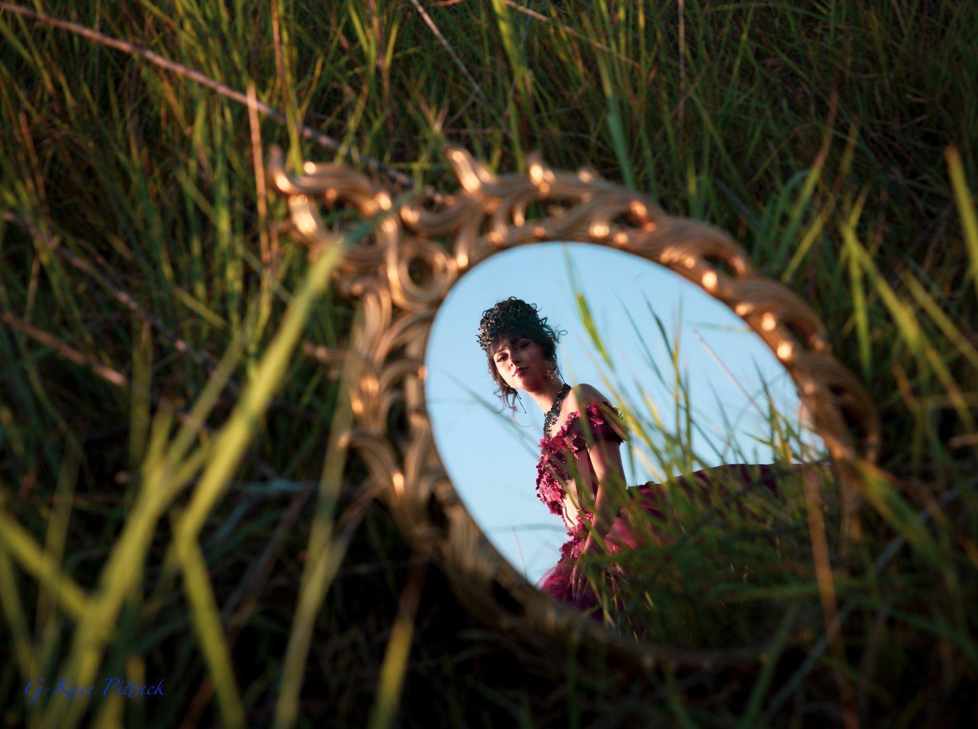 Mirror reflecting a woman in a pink dress in tall grass with a blue sky background.