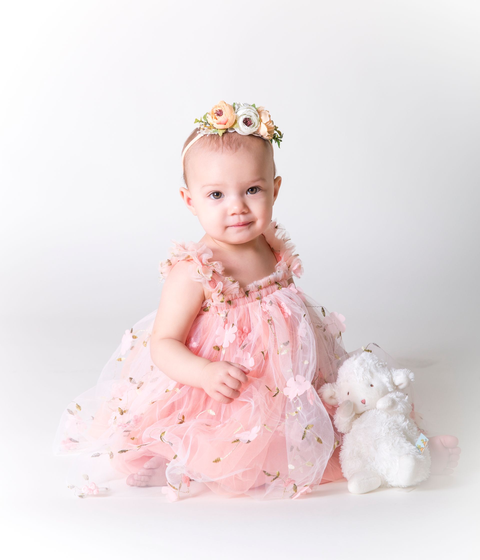 Baby in pink floral dress and headband, sitting with a white stuffed animal, smiles.