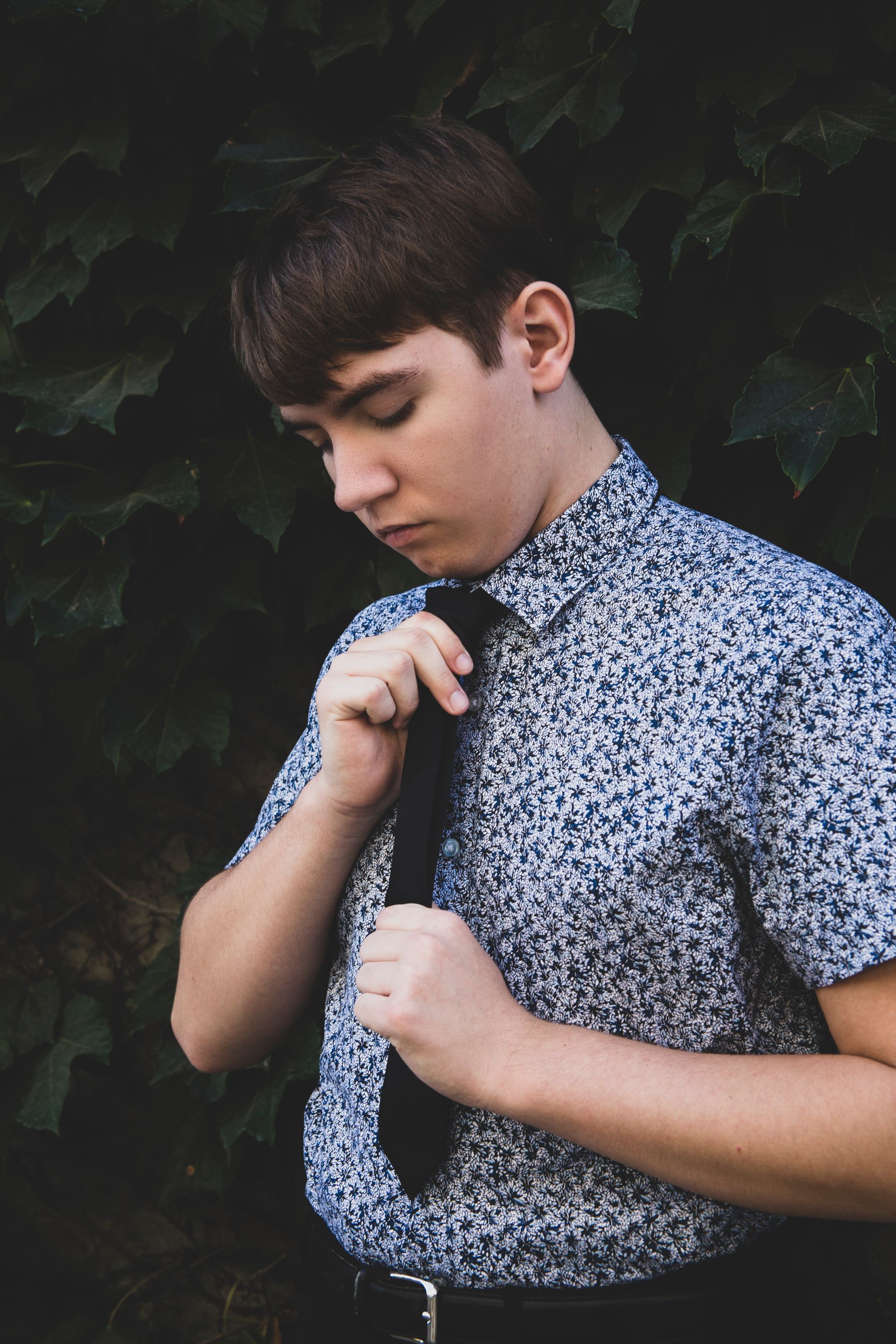 Young man in patterned shirt and tie, adjusting tie, in front of dark green foliage.