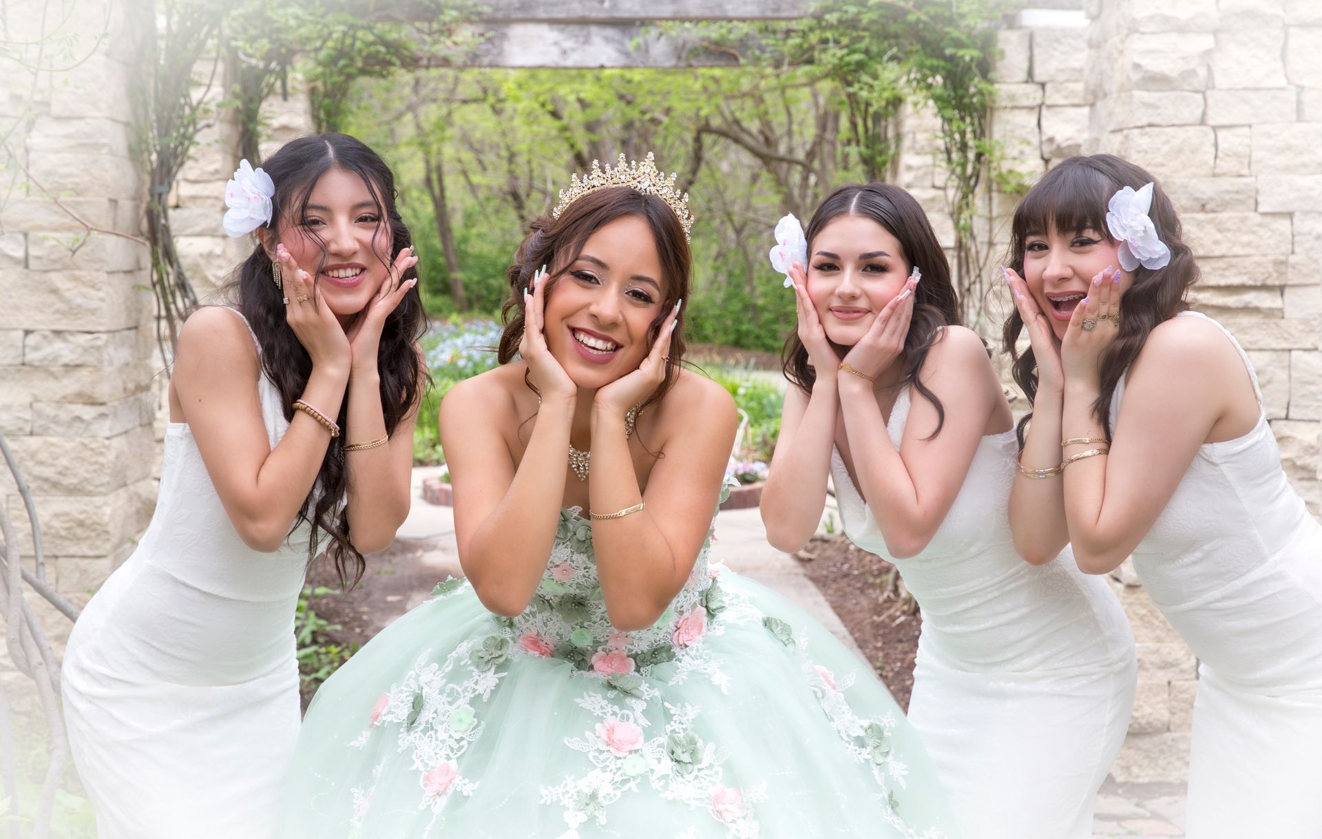 Four women posing for a photo in front of stone archway. The woman in the center wears a ballgown, and the others wear white dresses.
