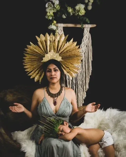 Woman in gold headdress, holding child, dark background with macrame.