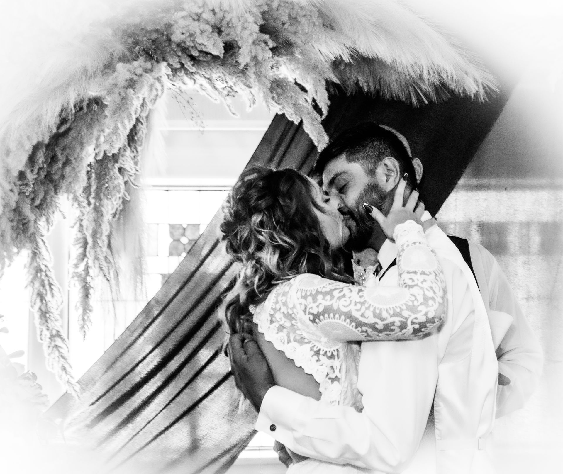 Couple kissing at a wedding ceremony, beneath a floral arch. Black and white photo.