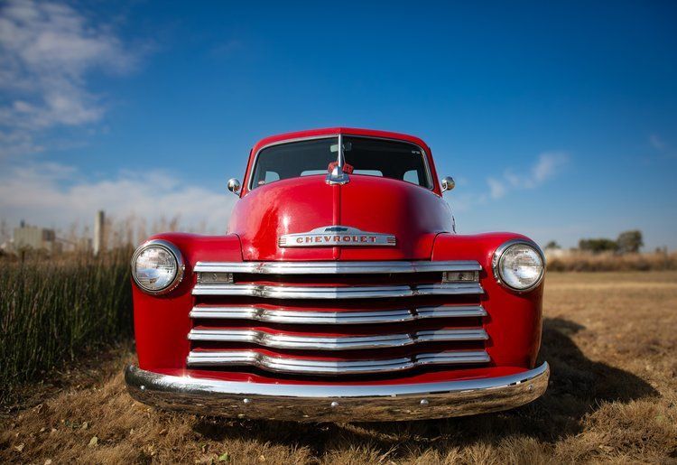 Red vintage Chevrolet pickup truck parked in a field with a blue sky in the background.