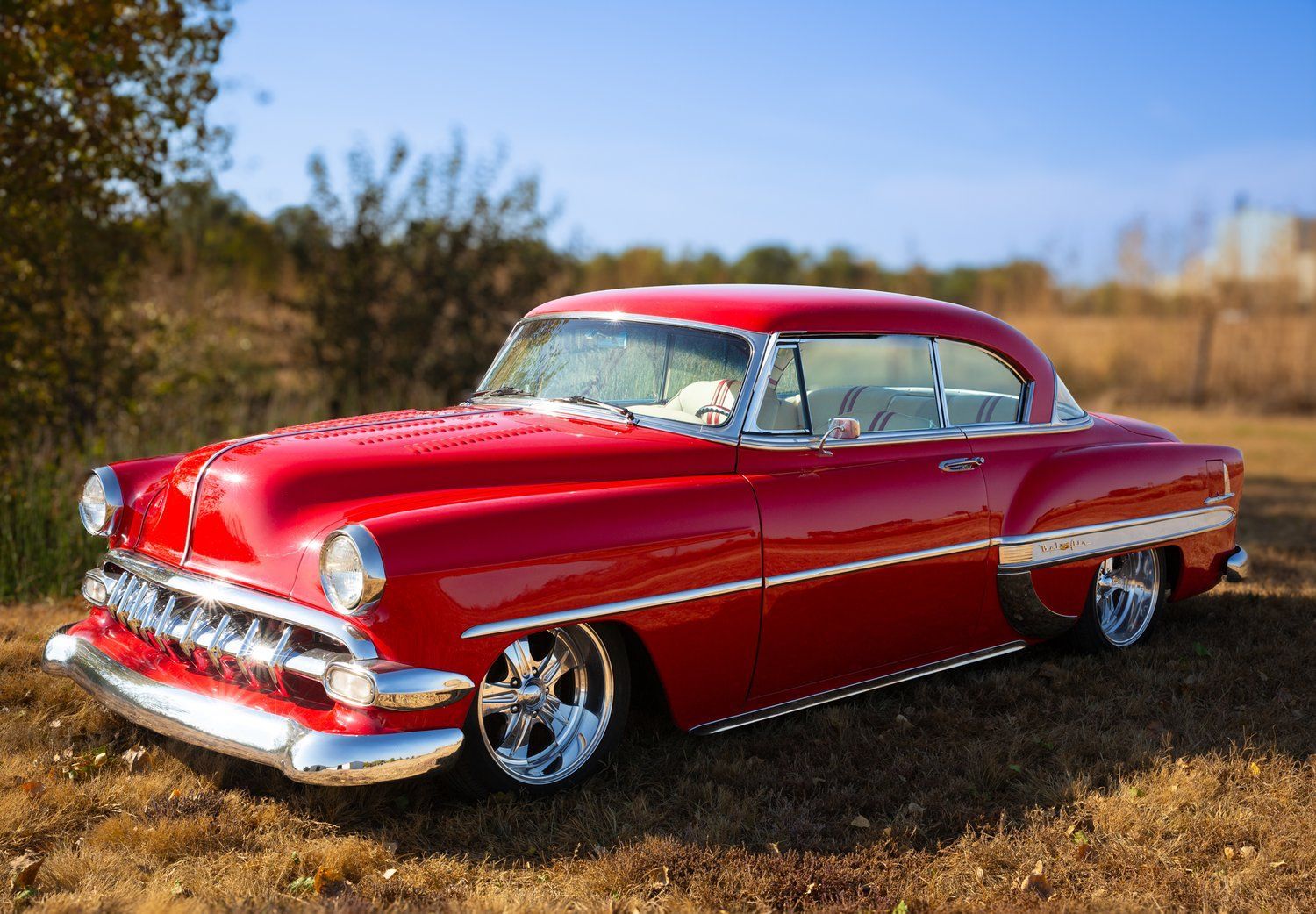 Red classic car with chrome accents parked in a grassy field on a sunny day.