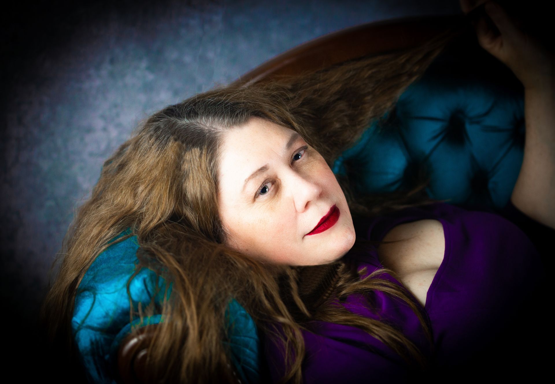Woman with long brown hair, red lipstick, wearing a purple top, lounging in a blue velvet chair.