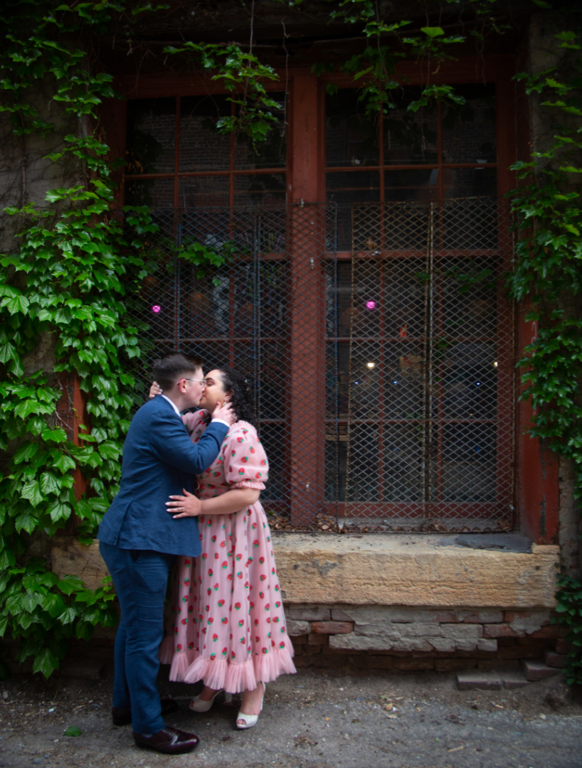 Couple kissing in front of a window with ivy, woman in pink dress, man in blue suit, outdoors.