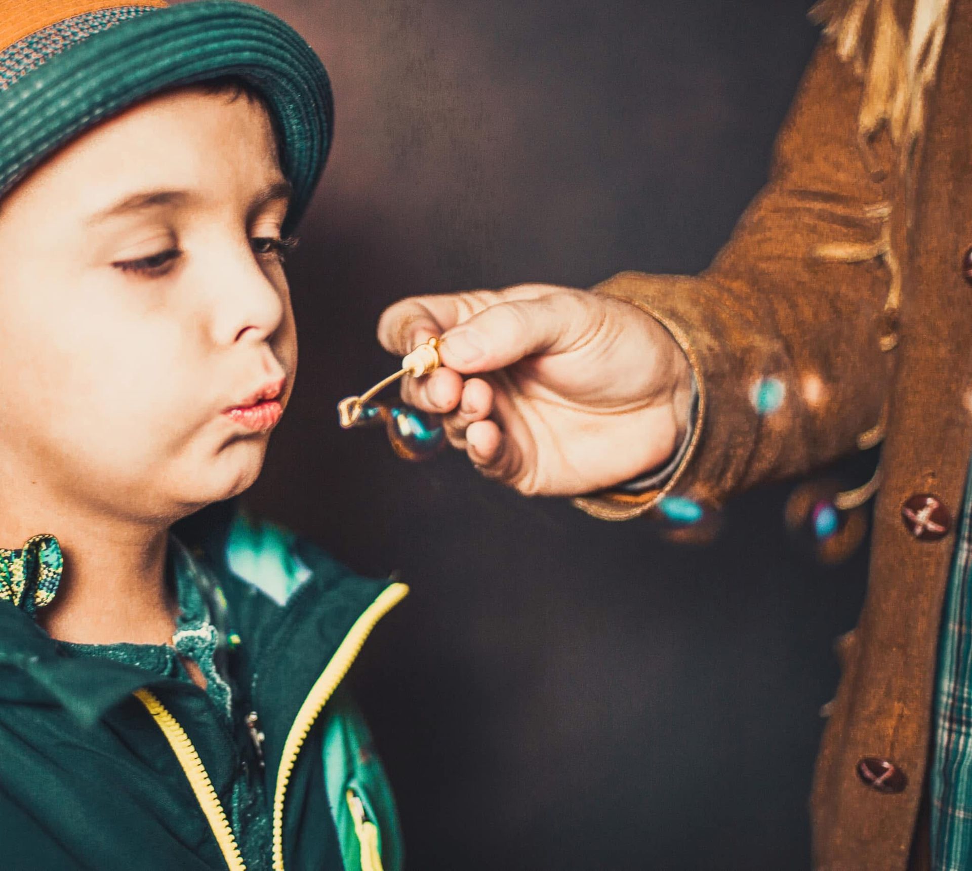 Boy in hat blowing bubbles held out by an adult.