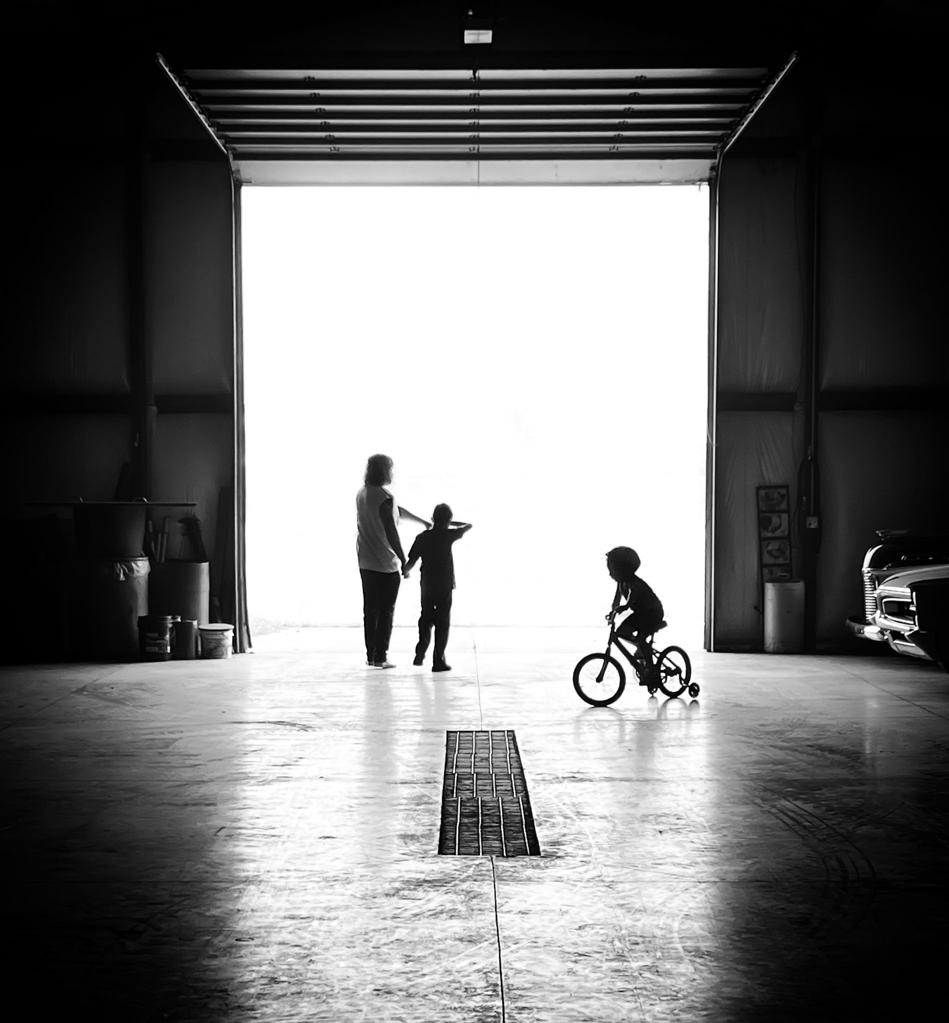 Family silhouetted in doorway, one child on a bike, the other two standing in a garage, bright light outside.