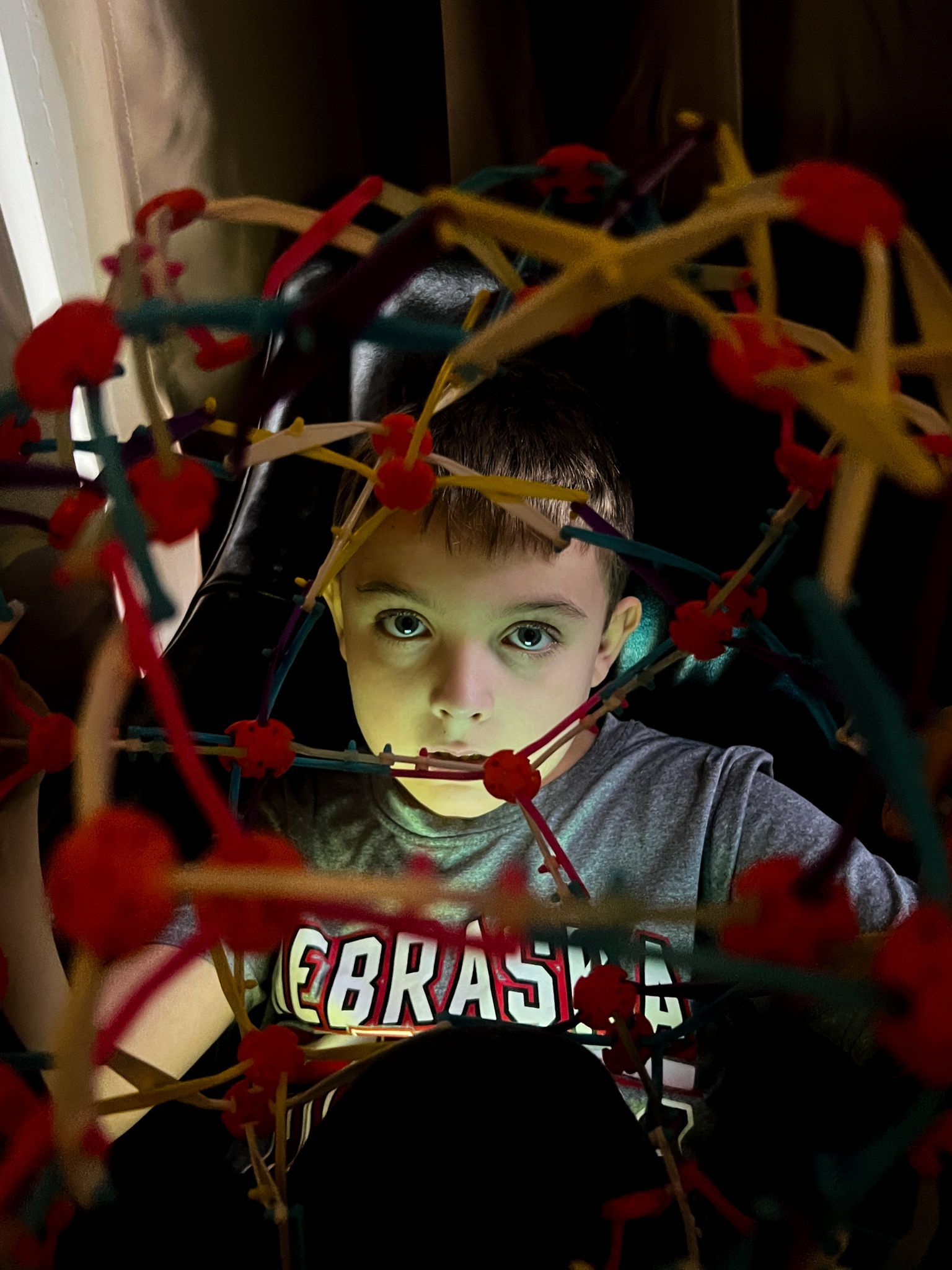 Boy looks through colorful pipe cleaner structure, lit from within.