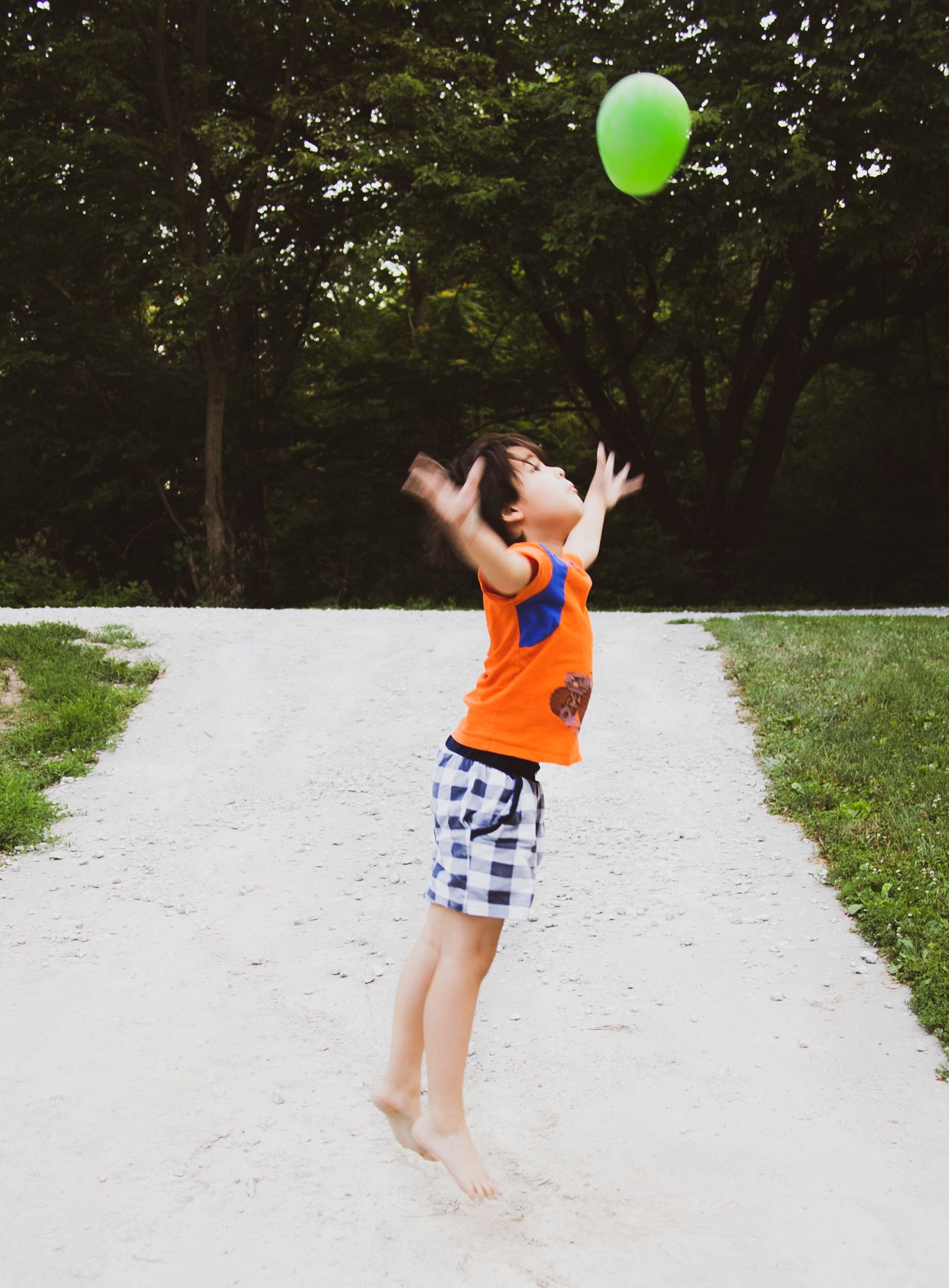 Boy jumping up to catch a green balloon on a white path, arms raised, in a park.