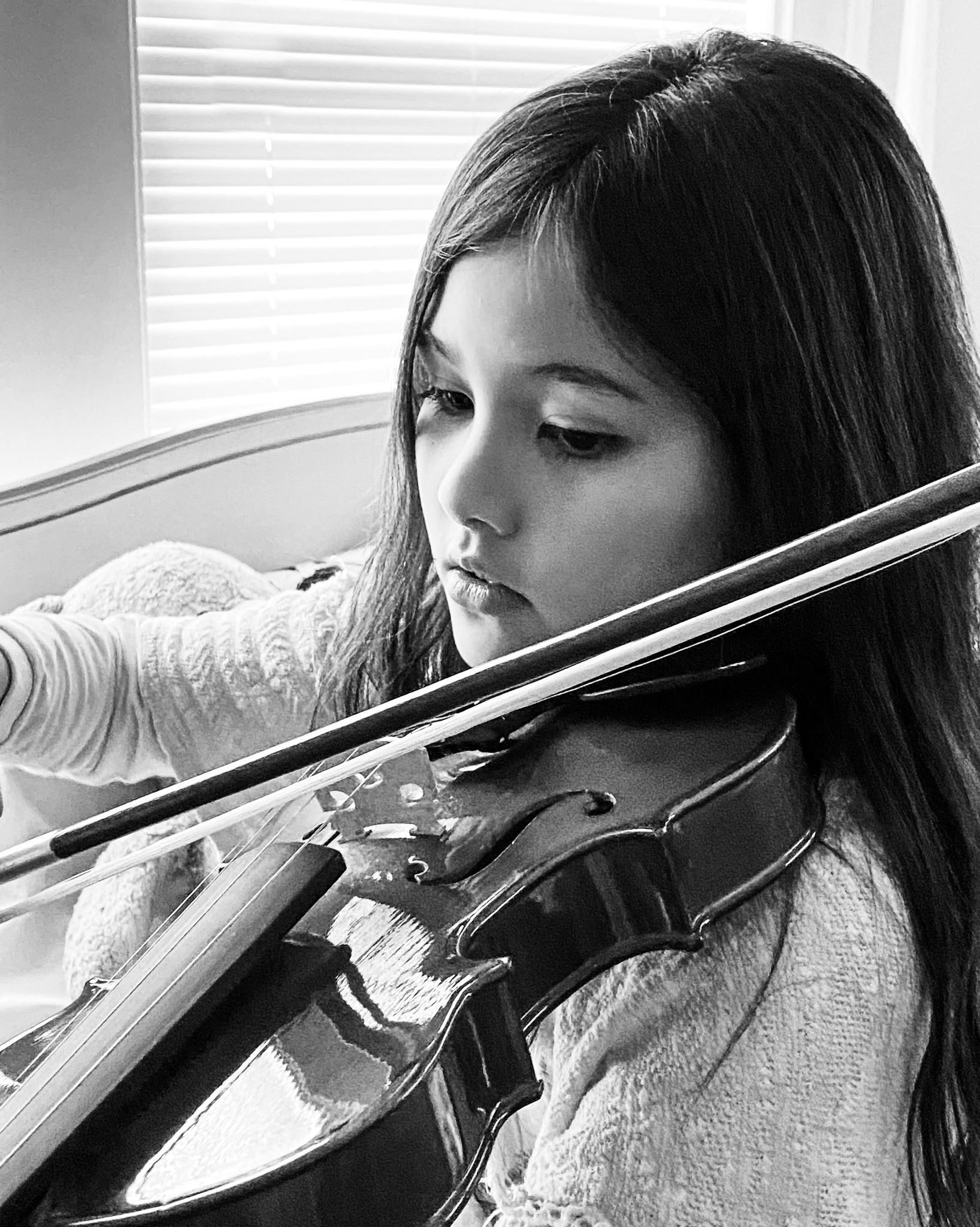 Girl playing the violin, looking focused. Indoors, near a window. Black and white photo.